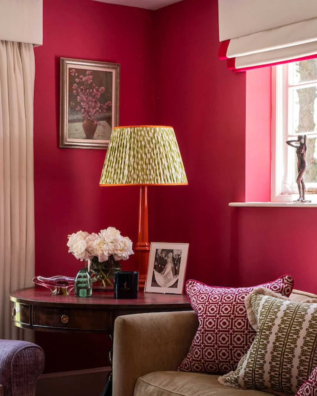A styled living room corner with deep red walls, a patterned green and gold pleated lampshade, a dark wood side table with a vase of white flowers, and matching red and cream curtains.